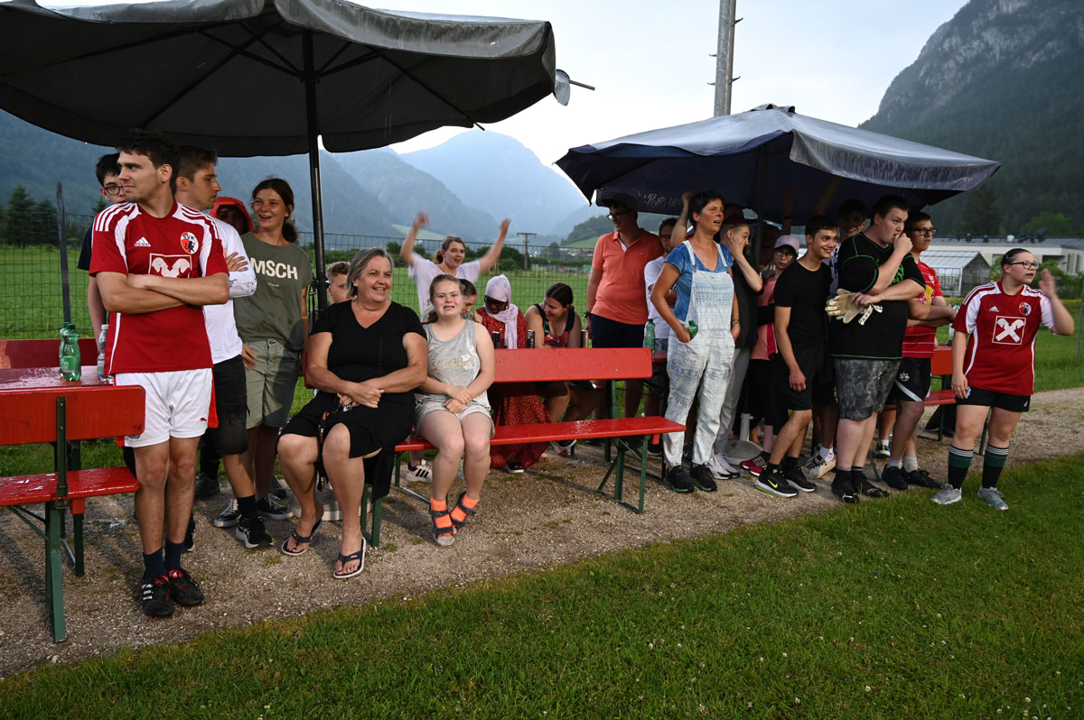 Fans beider Teams sitzen unter Schirmen am Spielfeldrand