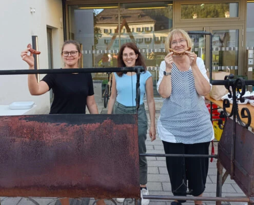 Zwei Wohnbetreuerinnen und die Psychologin stehen hinter dem Grill am Campus von oberrainanderskompetent und zeigen fertige Würstchen.