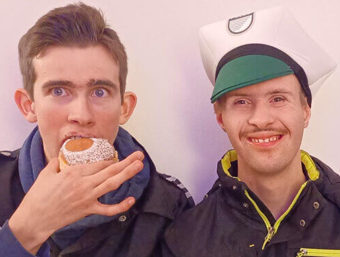 Zwei junge Männer mit Kostüm und Krapfen, fröhlich bei inklusiver Faschingsfeier in Salzburg