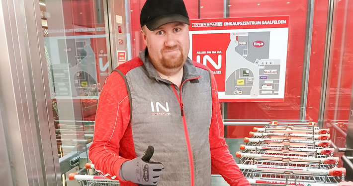 Mann mit Arbeitskleidung zeigt Daumen hoch vor Einkaufswagen