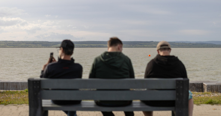 Drei Personen sitzen auf Bank am Seeufer