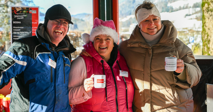 Drei Personen an einem Wintermarktstand lächeln und halten Becher